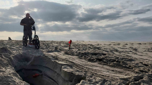 Man standing on a beach employs a ground based radar machine on the sand. 