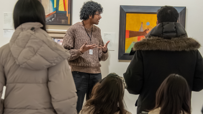 Group of people watch a tour guide discuss an artwork on the wall of a gallery.