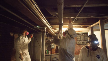 In a dimly lit basement. three volunteers in white coveralls and respiratory masks work, with large brushes, to remove mold from the ceiling. January, 2013: Volunteers from from the Respond and Rebuild — a disaster response collective that came together to the Rockaway Community after Sandy — remove mold from a house damaged by Hurricane Sandy.