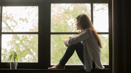 thoughtful girl sitting on sill embracing knees looking at window