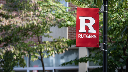 A red banner with the Rutgers block R logo hangs on a lamp post on campus against a background of green trees
