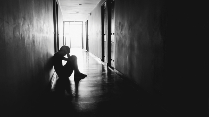 A black and white photo showing a person in a seated crouch against the wall of a long hallway, silhouetted by light from a far doorway.