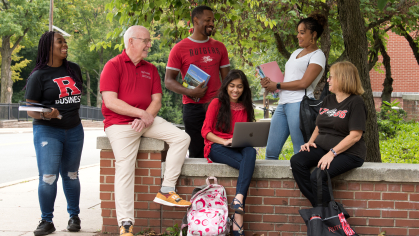 A diverse group of individuals wearing Rutgers apparel sitting on and standing around a short brick wall