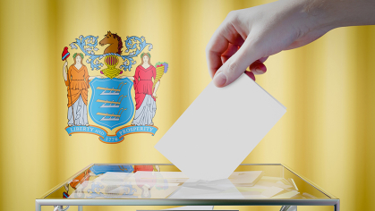 A person putting a slip of paper in a ballot box in front of the flag of New Jersey