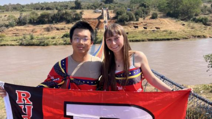 Two students stand on a bridge overlooking river, holding a Rutgers R flag together