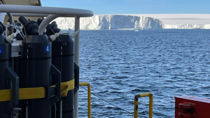 Sampling rosette with gray sampling bottles at left, the ship’s rail at lower right, and the face of the ice shelf in the background.