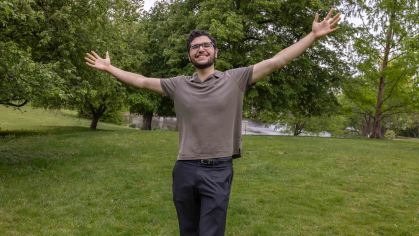 Rutgers junior Adnan Alkhalili is smiling as he raises his arms and stands in grass near Passion Puddle between the Cook and Douglass campuses.