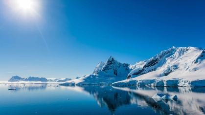 A set of icy mountains at right and moutains in the horizon to the left with a blue glassy lake in the foreground