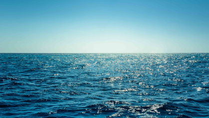 A wide expanse of blue ocean with a medium blue sky with brightness at the horizon, suggesting a sunrise.