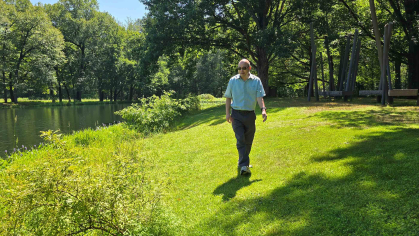 Man walks along wooded lawn with a body of water to his left.