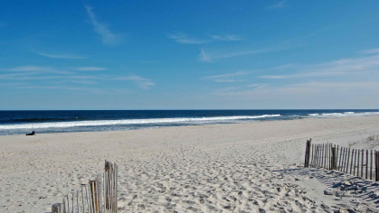 A large white-sand beach with waves lapping at the shore. A dune fence is in the foreground.