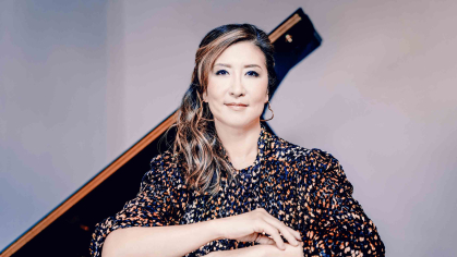 Rutgers professor Min Kwon is in seated pose, with her forearms resting on a black shine table surface with a raised piano lid in the background.