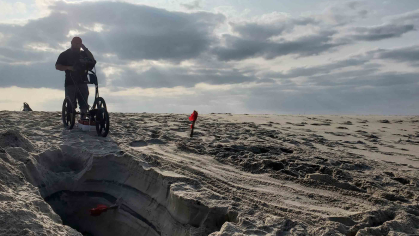 Man standing on a beach employs a ground based radar machine on the sand. 
