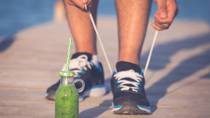 A close-up of someone's legs below their knees. The person is tying one of his sneakers and has placed a bottle of green juice in front of him.