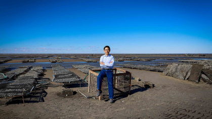 Man sits on an oyster crate on beach sand flats with the horizon of sea behind him