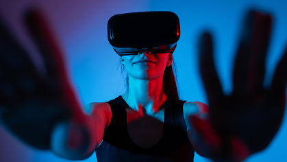 A woman in a dark room with a blue background reaches forward while wearing a virtual reality headset