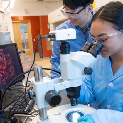 Two students working together looking into a microscope with a computer screen