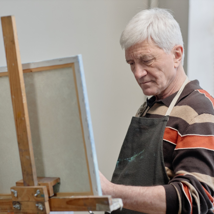 Senior citizen man painting on canvas in classroom setting, standing at easel and focusing on artwork.