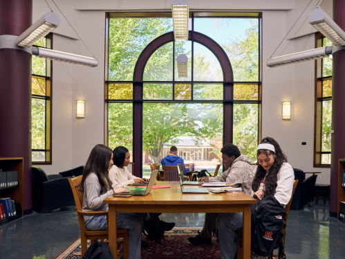 A group of students, Alyson Frio, Tiara Clyde, Jean Laurard, and Yasmeen Higazy, studying in the library in front of a large window