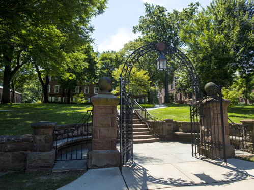 Green trees cover the grounds of  Old Queens as seen through the Class of 1902 Memorial Gateway on Hamilton Street