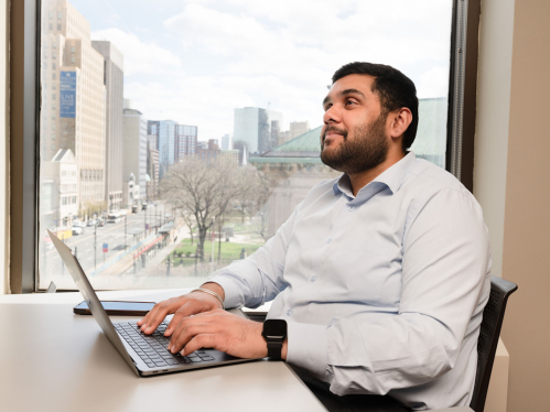 A student works on a laptop in front of a window with a view of the city 