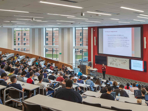 A classroom with stadium seating filled with students with a professor at center teaching