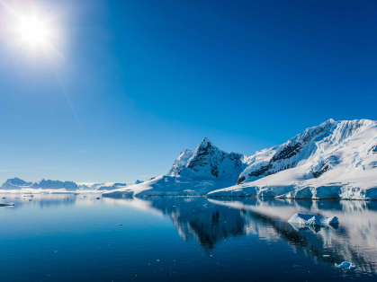 A set of icy mountains at right and moutains in the horizon to the left with a blue glassy lake in the foreground
