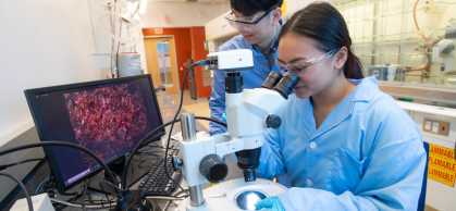 two students in lab coats with a microscope and computer screen