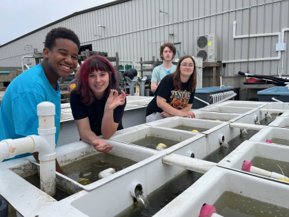 Four people posing as they stand in-between rows of square containers. The person second from the left holds a handful of small oysters in one hand and a singular small oyster pinched between fingers in her other hand.