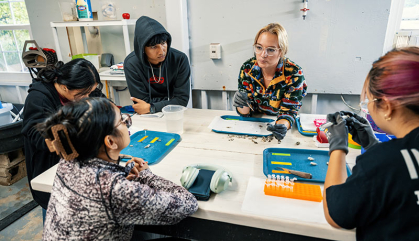 A group of students at a lab table listen to a scientist.