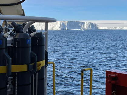 Sampling rosette with gray sampling bottles at left, the ship’s rail at lower right, and the face of the ice shelf in the background.