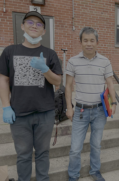 Two men stand in front of a brick building.