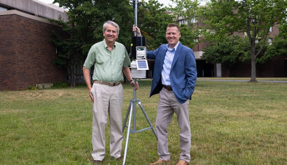 Two men stand outside next to weather equipment.