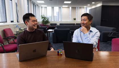 Two men set at a table with their laptops in front of them. The men are talking.