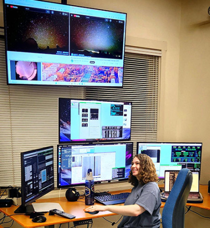 A woman sits at a desk surrounded by screens with colorful data displays.