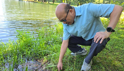 Man in blue shirt crouches by a body of water examining an embankment.