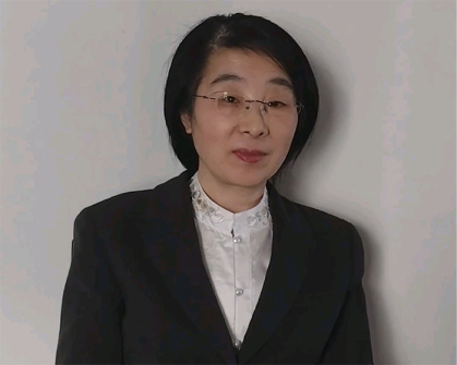 Woman against a white wall. She is wearing a black jacket and a high-necked white shirt.