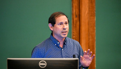 Man in blue shirt speaks with a green chalkboard behind him.