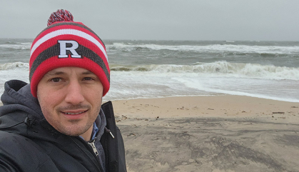 Man in a red Rutgers cap stands on a beach with waves in the background.