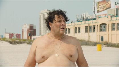 A large, bare-chested man stands on a beach in a scene from "The Clam Guy."
