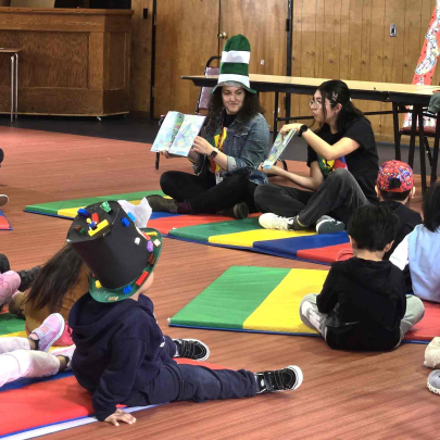 Two young women sitting on gym floor read a children's book in Spanish and English to young children.